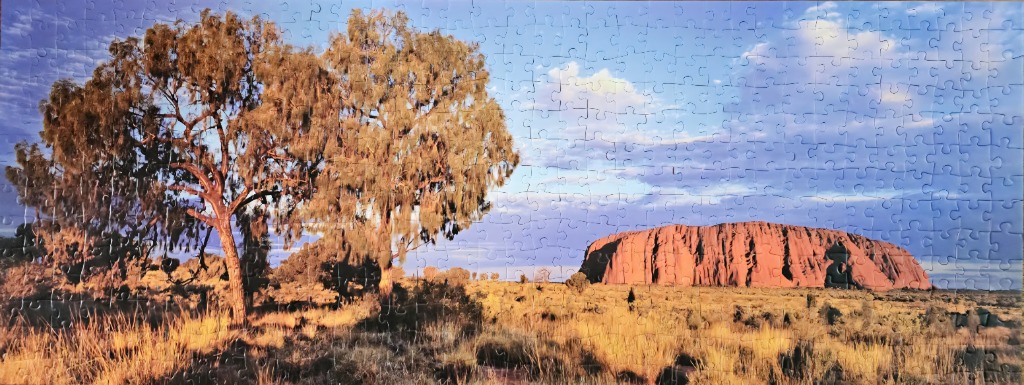 A panoramic photograph of Uluru at sunset with desert trees in the foreground.