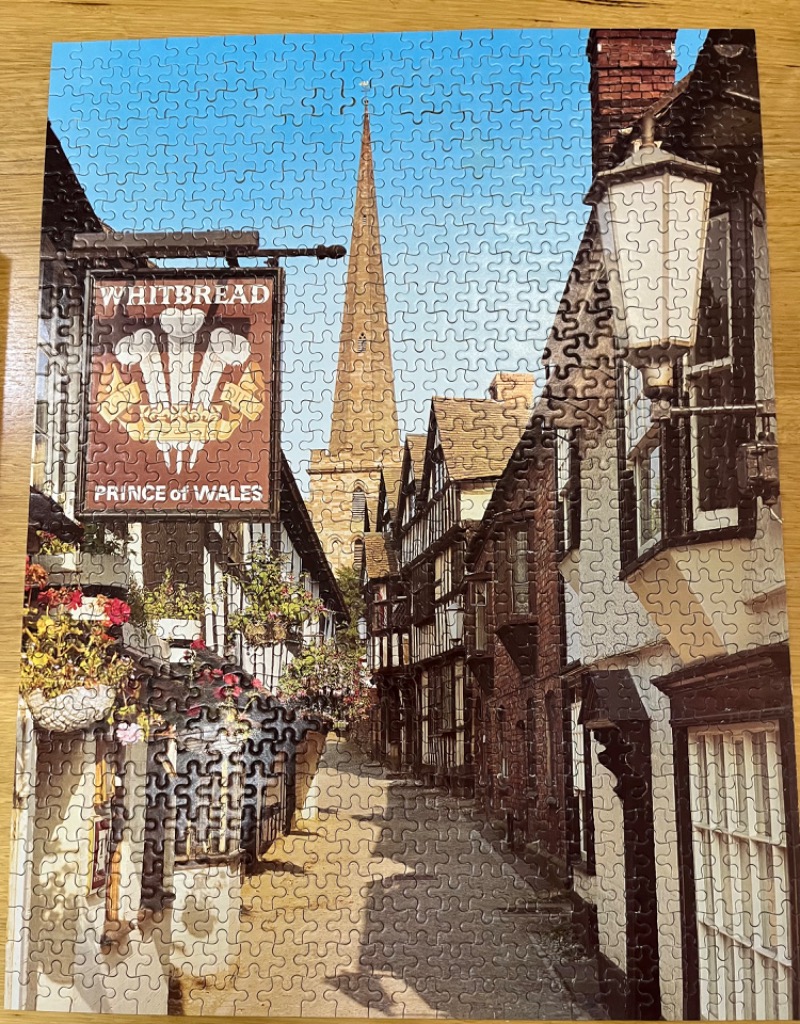 A narrow historic English street leading to a tall stone church spire, with a hanging Prince of Wales pub sign in the front.
