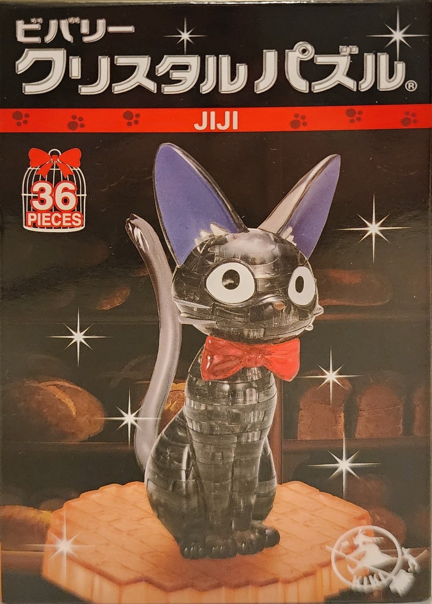 A black crystal cat figurine with a red bow tie sitting on an orange platform against a dark bakery background.