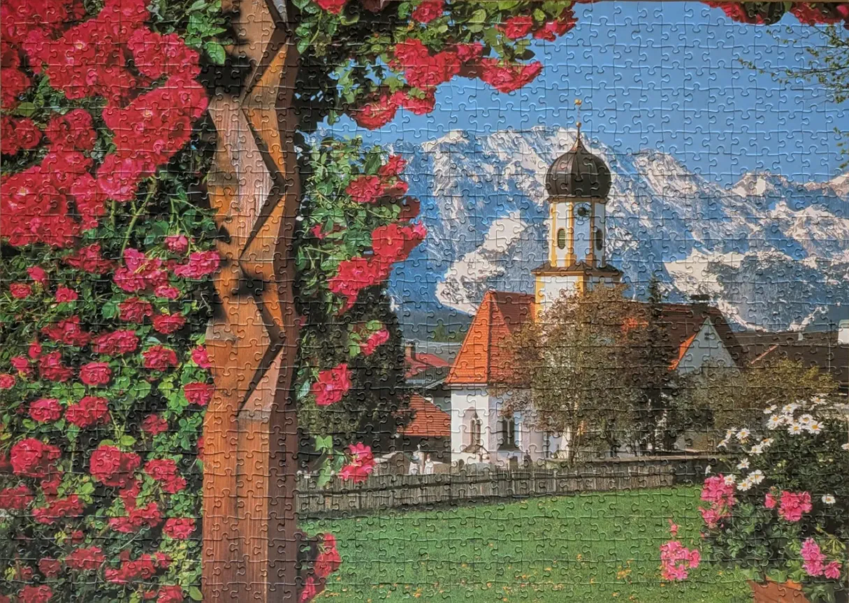 A scenic view of a church with a red roof, a wooden trellis covered in pink roses, and snow-capped mountains in the back.