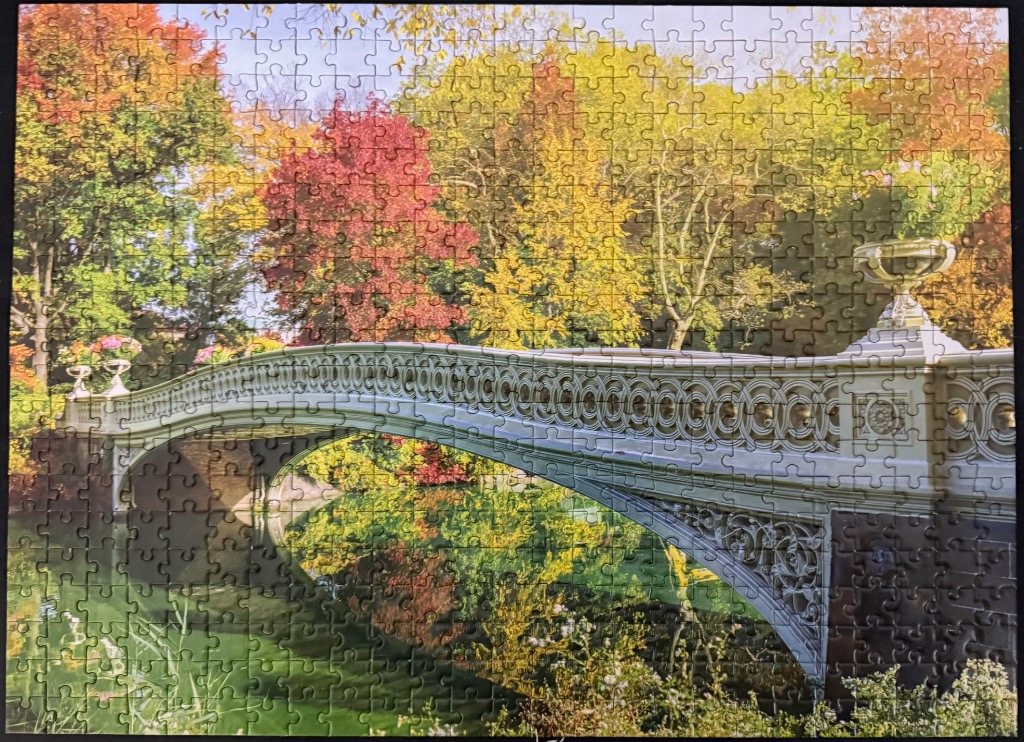 A photograph of Bow Bridge in Central Park during autumn, with vibrant yellow and orange trees reflecting in the water.