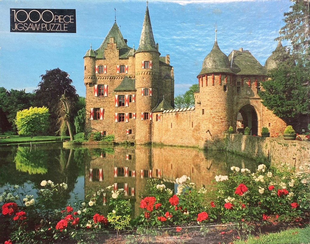 A stone castle with red and white shutters reflected in a moat, surrounded by red and white roses in a garden.