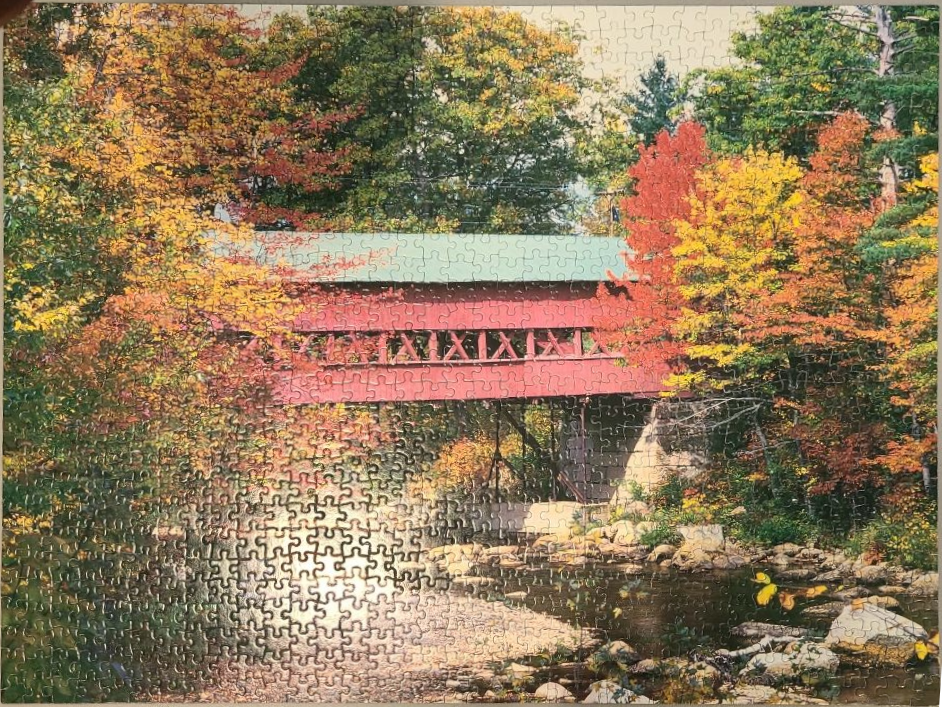 A red covered bridge spanning a shallow creek surrounded by vibrant autumn trees.