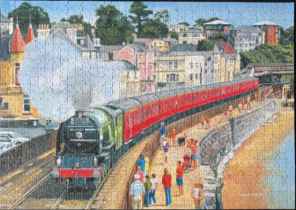 A green steam engine pulling red carriages past a seaside promenade with people and a town in the background.