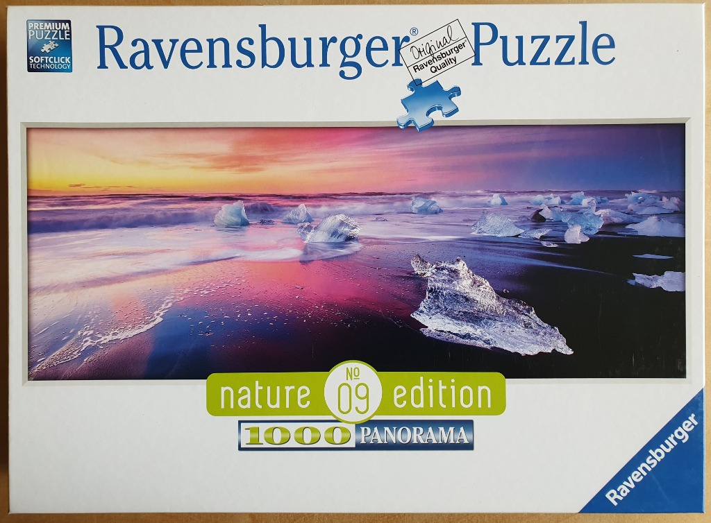 A panoramic photograph of icebergs on a black sand beach at sunrise or sunset with vibrant pink and gold skies.