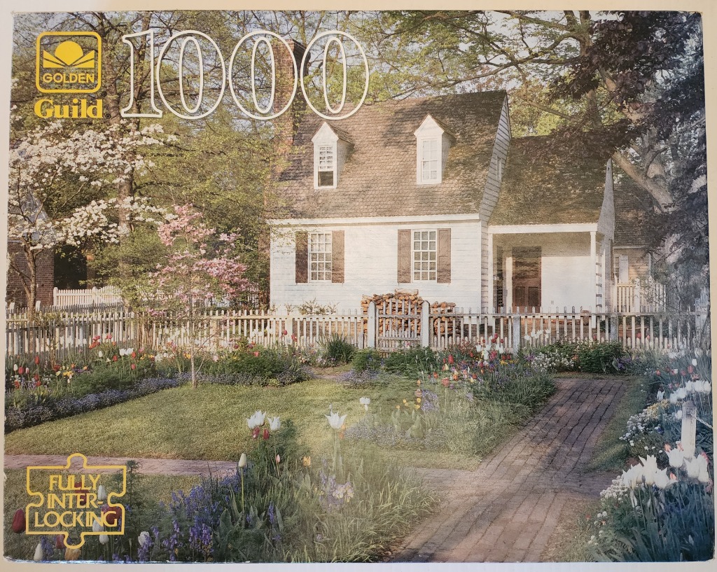 A white colonial cottage behind a flower-filled garden and a white picket fence in spring.