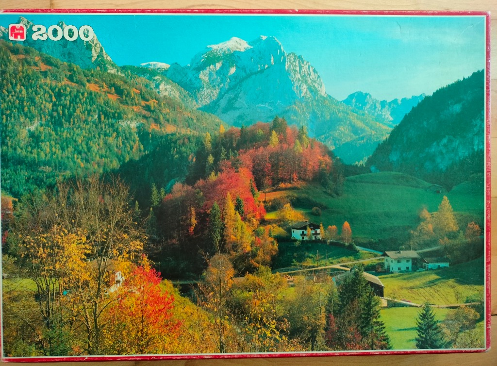 An autumnal mountain landscape featuring rugged snowy peaks, forested slopes, and quaint valley houses.