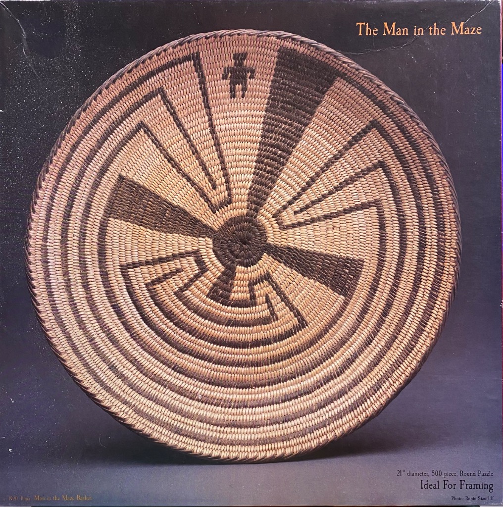 A close-up photograph of a round, woven basket featuring the traditional Man in the Maze pattern against a dark background.