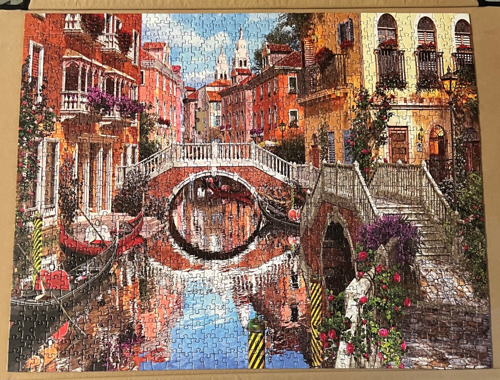 A romantic painting of a sunlit Venice canal with stone bridges, gondolas, and old buildings with flower-filled balconies.