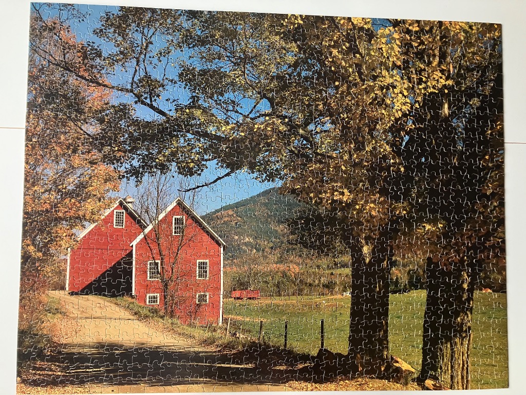 A rural landscape featuring a pair of red barns nestled among autumn trees in front of a mountain.