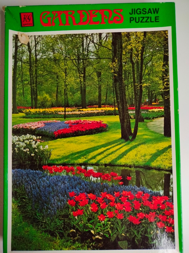 A lush spring garden featuring vibrant red and blue flower beds along a pond with tall trees in the background.