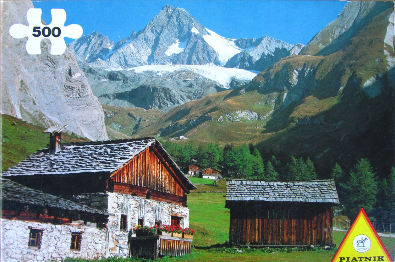 A sunny mountain landscape with wooden alpine huts in a green valley beneath snow-capped peaks.