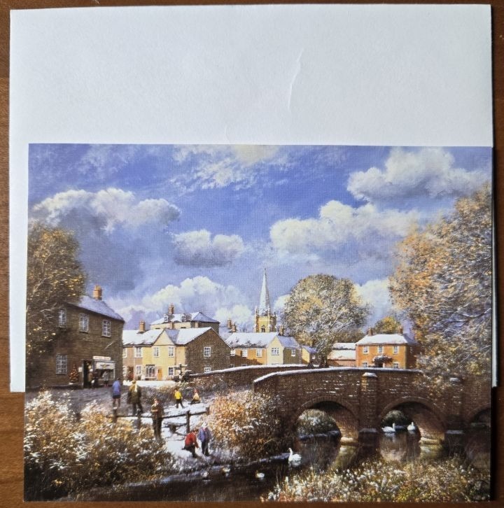 A wintry English village scene featuring a stone bridge, historic homes, and a church spire under a cloudy blue sky.