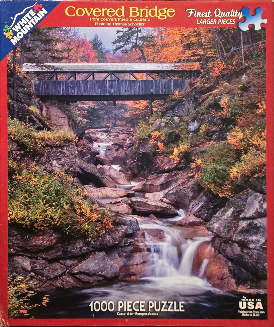 A wooden covered bridge spans a rocky gorge filled with a cascading stream surrounded by autumn trees.