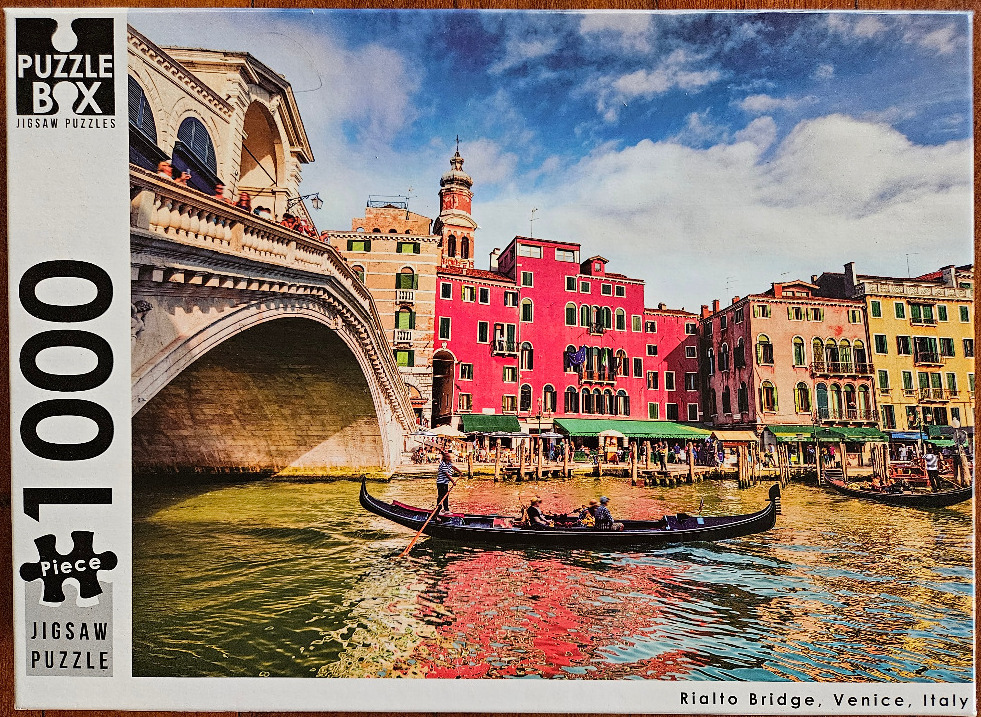 A sunny day scene of the Rialto Bridge over a canal in Venice, Italy with a gondola in the foreground.
