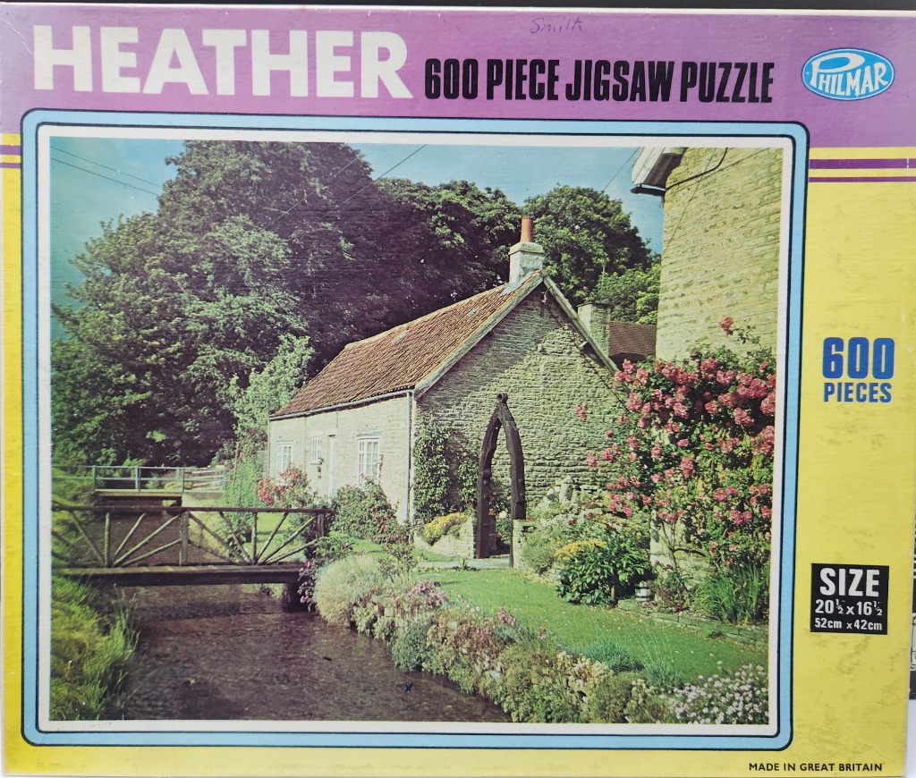 A stone cottage beside a small stream with a footbridge and blooming pink rose bushes.