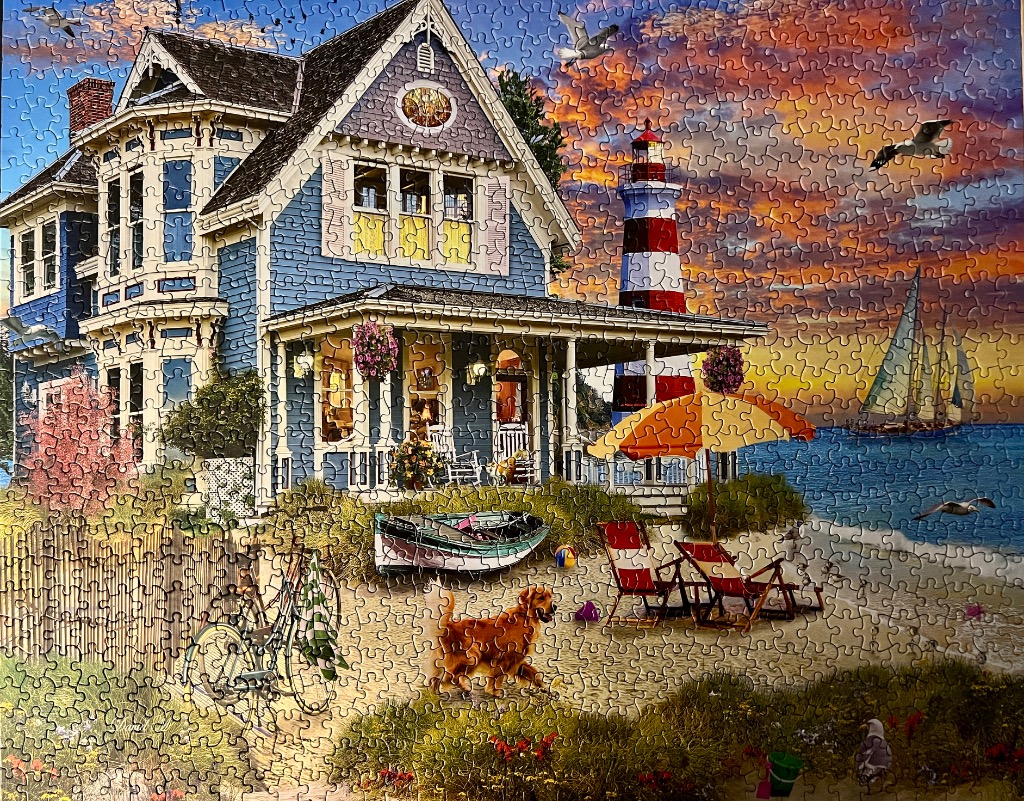 A blue Victorian house by the seaside at sunset with a lighthouse, sailboat, and golden retriever on the beach.