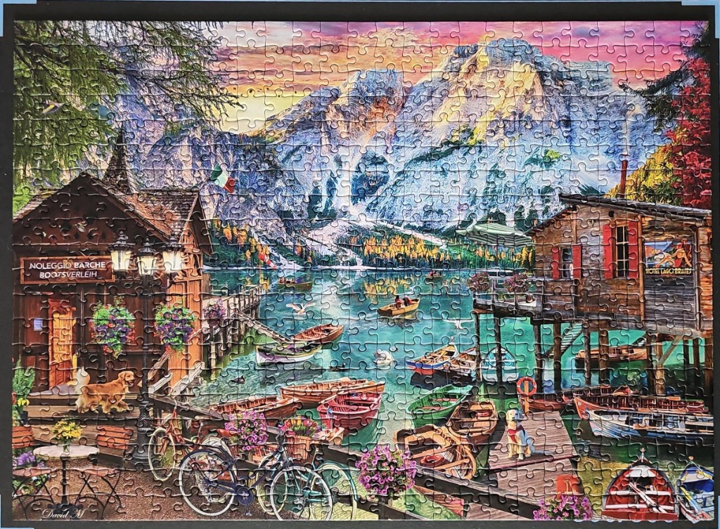 A peaceful lakeside scene with wooden boathouses, boats, and a mountain backdrop at sunset.