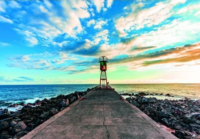 A lone metal beacon stands at the end of a stone pier extending into a calm ocean under a vibrant, multi-colored sky.