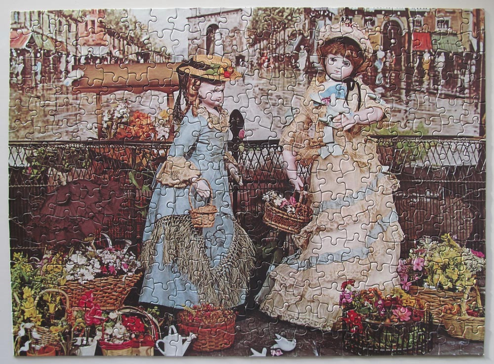 Two vintage-style porcelain dolls in period dresses standing in an outdoor flower market.