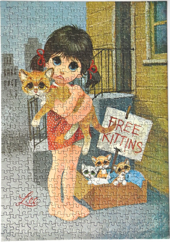 A wistful girl with large eyes holding a kitten next to a cardboard box of kittens with a sign that says FREE KITTINS
