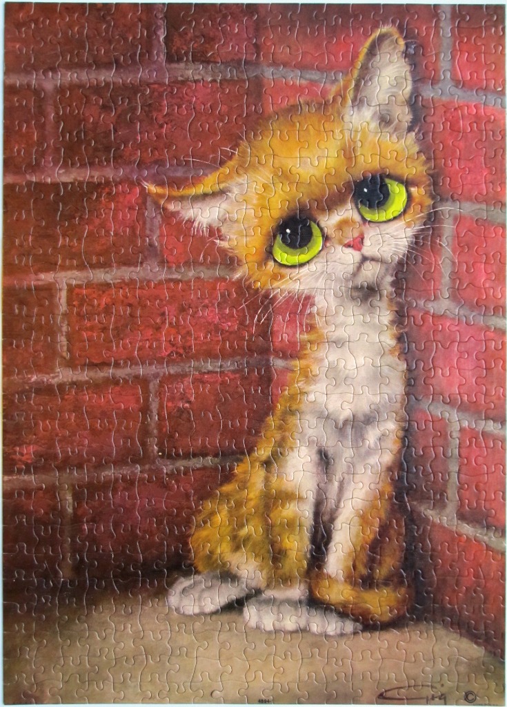 A wide-eyed orange tabby kitten sitting against a red brick wall.