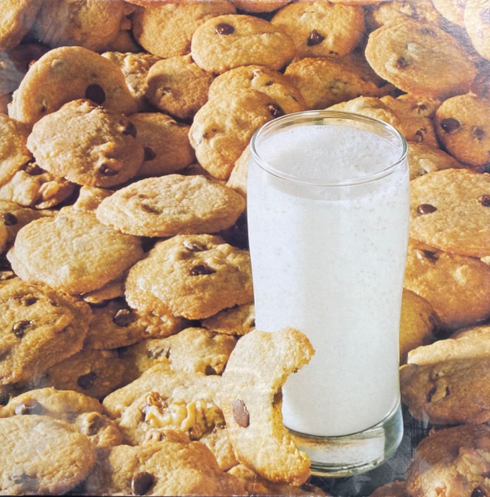 A tall glass of milk centered in front of a dense pile of golden-brown chocolate chip cookies.