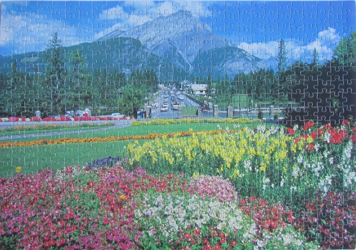 A vibrant flower garden in the foreground with a town bridge and mountain backdrop under a blue sky.