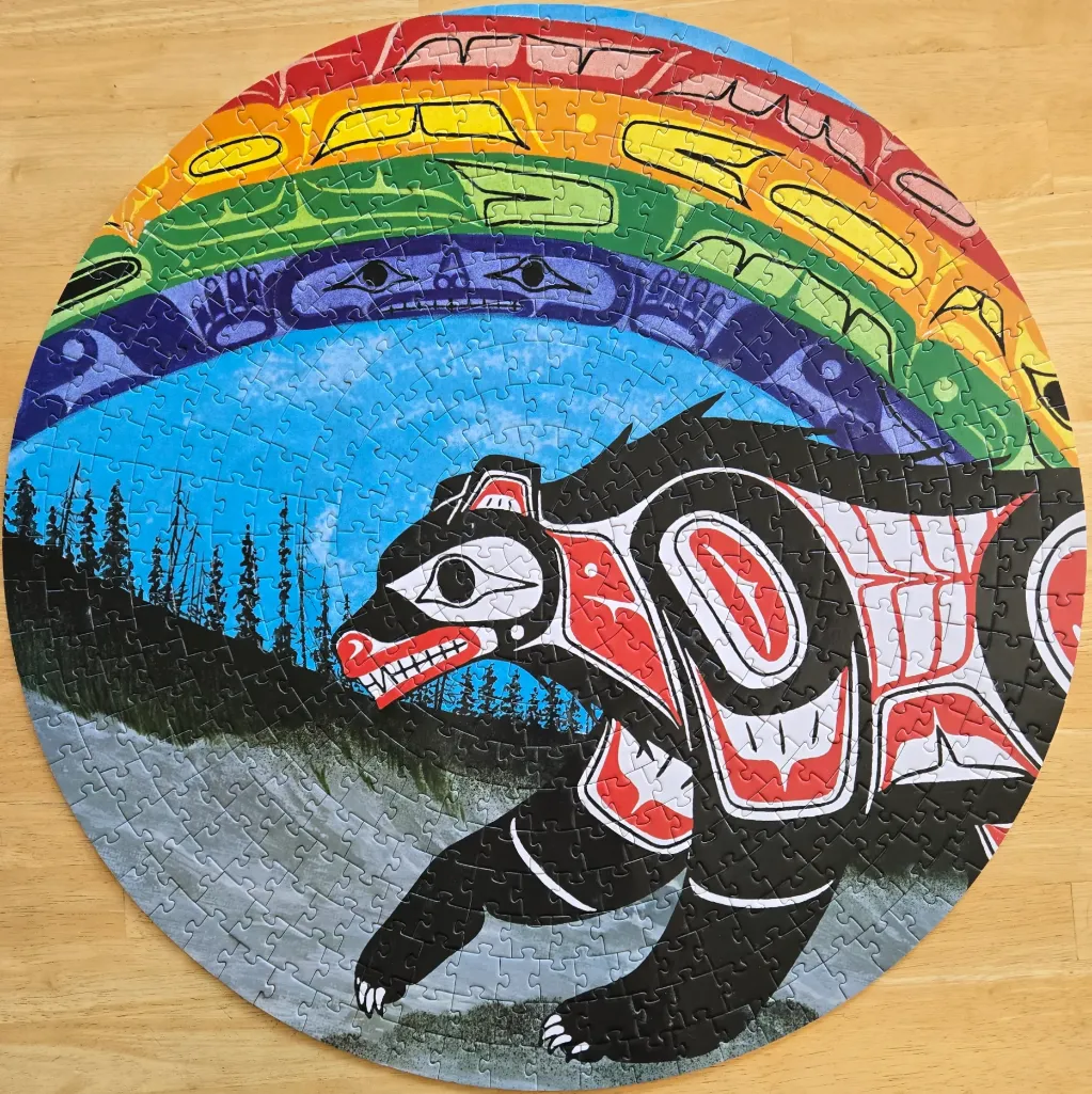 A circular artwork featuring a stylized bear in Pacific Northwest Indigenous art style under a rainbow sun.