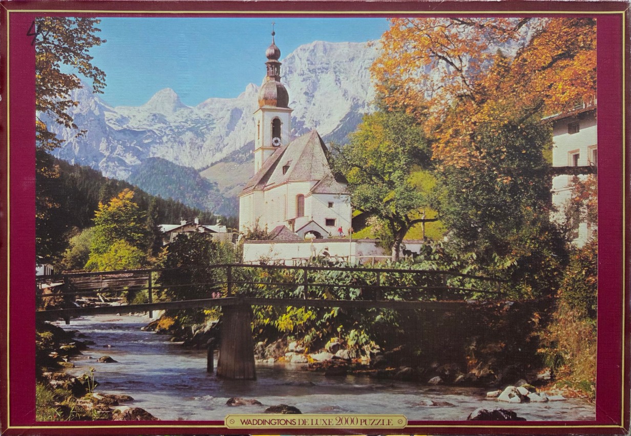 A scenic view of a white alpine church with an onion dome spire, a wooden bridge, and a river set against high mountains.