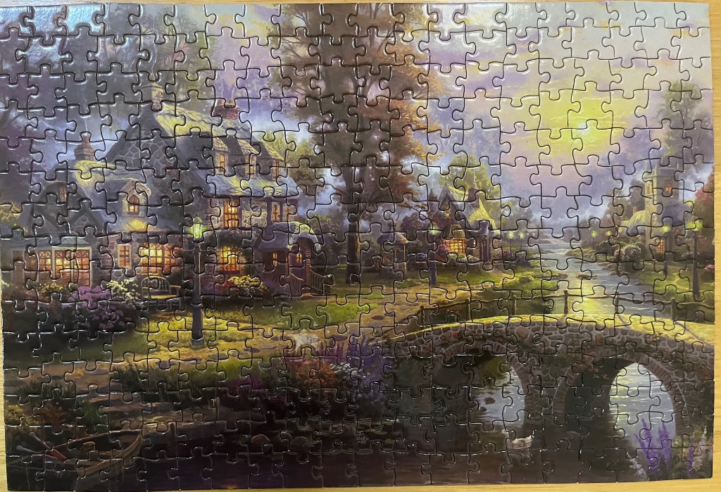 A scenic painting of a quaint stone cottage beside a stream with a stone bridge at sunset.