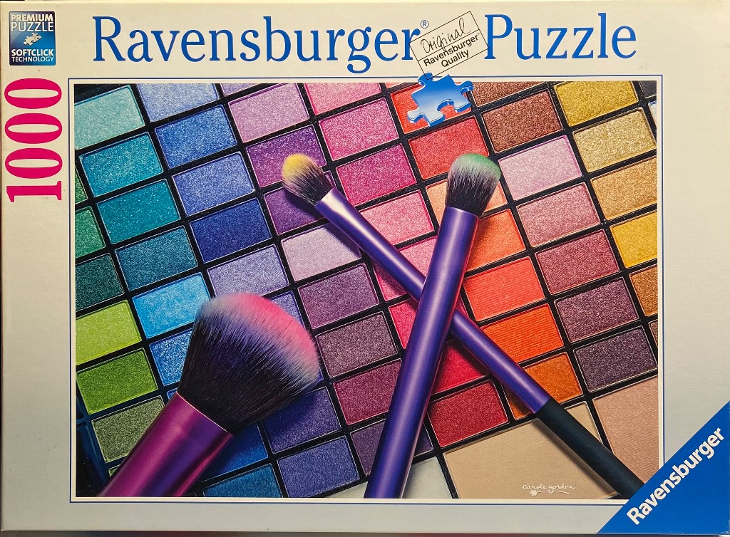 A close-up photograph of a vibrant makeup eyeshadow palette with two purple makeup brushes resting on top.