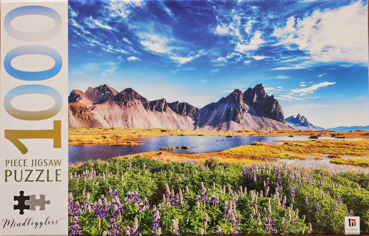 A panoramic landscape featuring a field of purple lupines in the foreground, a still lake, and jagged mountains in Iceland.