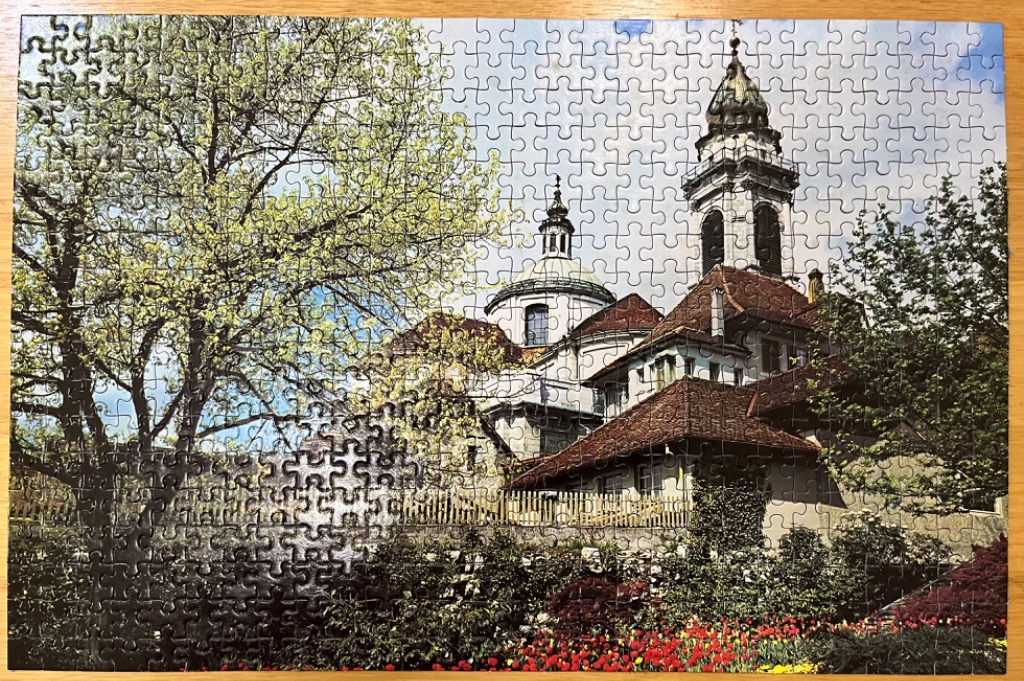 A sunny European cathedral with a dome and tower, framed by a large tree and a vibrant foreground of tulips.