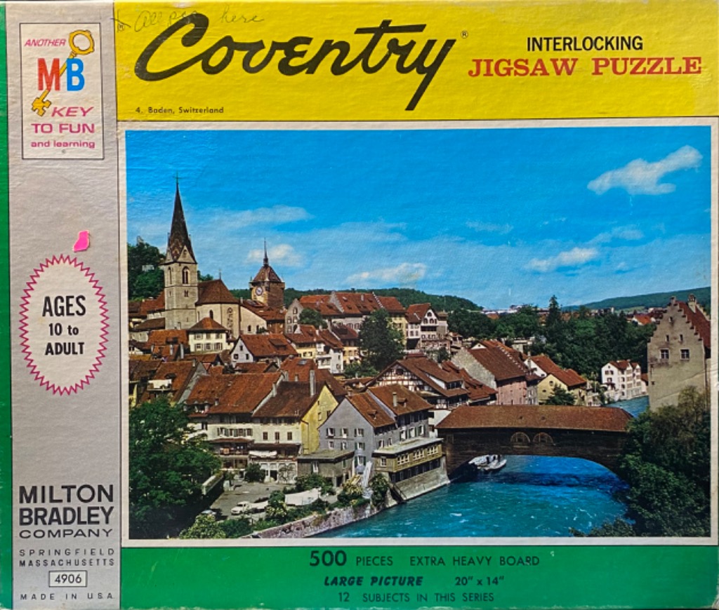 A scenic view of the Swiss town of Baden with a covered wooden bridge over a blue river and a church steeple.