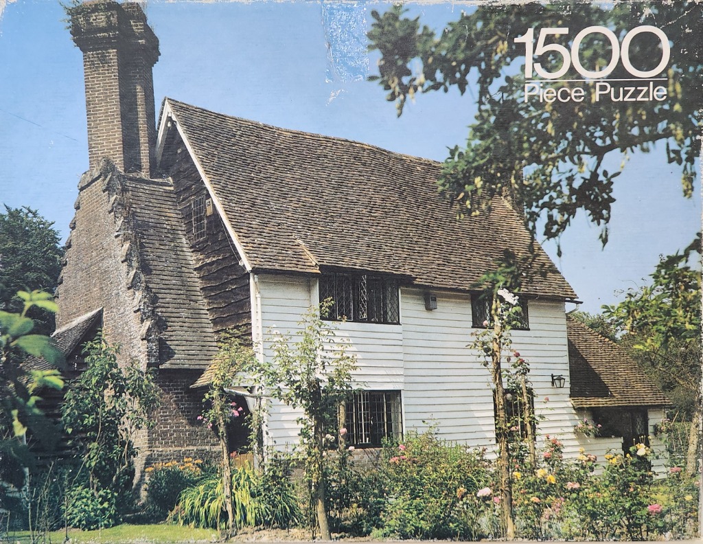 A historic white timber-framed cottage with a tall brick chimney, surrounded by a lush garden under a clear blue sky.
