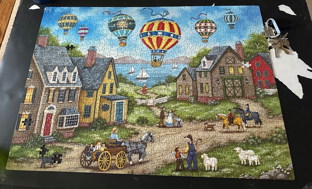 Folk art illustration of a colonial village with hot air balloons over a bay.