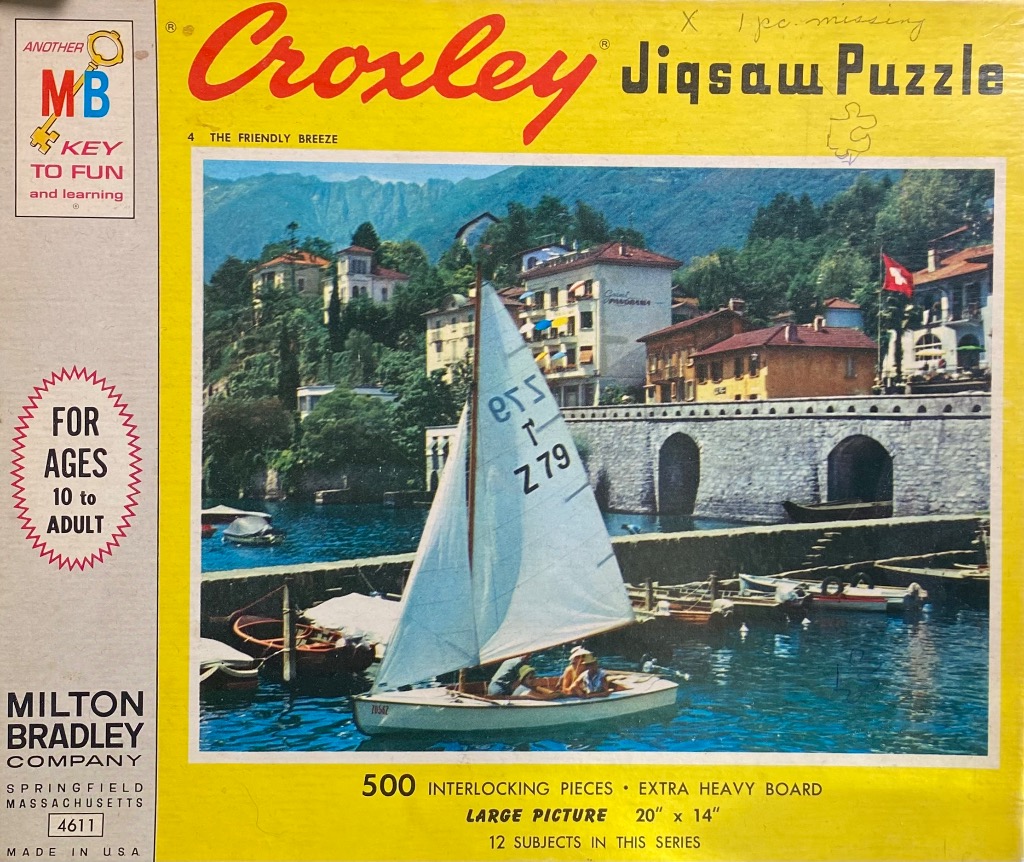 A sailboat gliding across a blue harbor in front of a European lakeside town with stone arches and mountains.