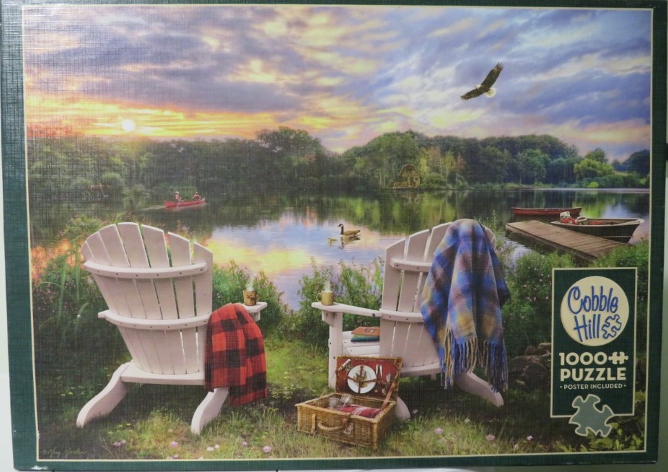 Two Adirondack chairs facing a serene lake at sunset with a picnic basket and blankets.