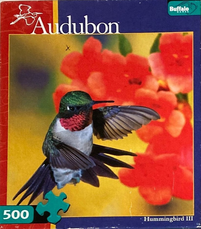 A hummingbird with iridescent green feathers and a bright red throat patch hovering in front of red flowers.