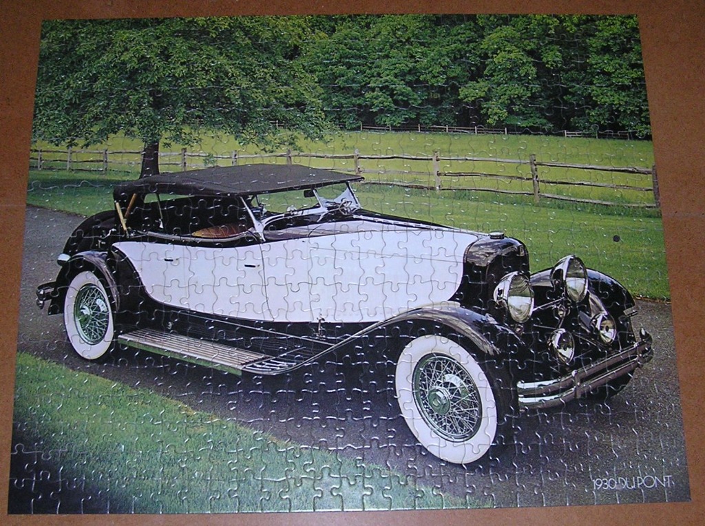 A vintage black and white 1930 DuPont automobile parked on a driveway with a green field and trees in the background.