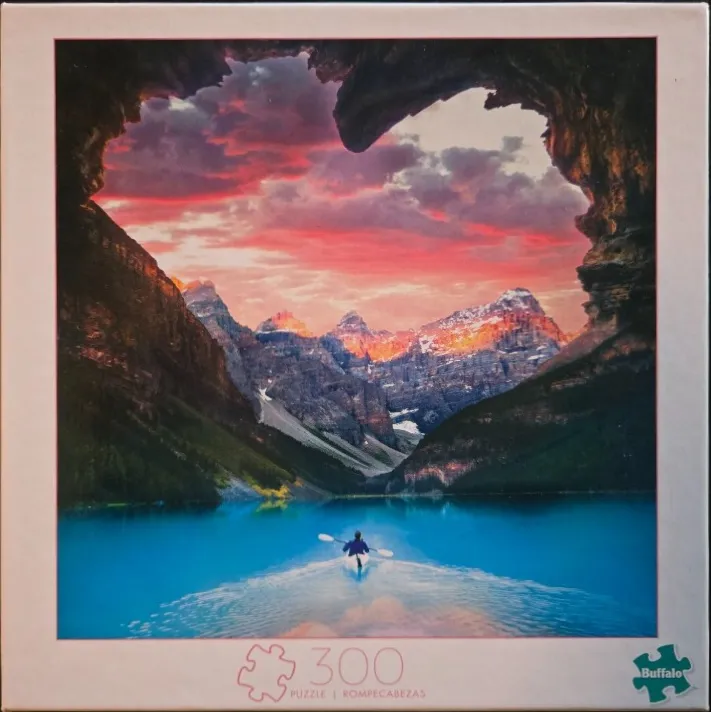 A kayaker on a turquoise mountain lake viewed through a heart-shaped rock cave opening at sunset.