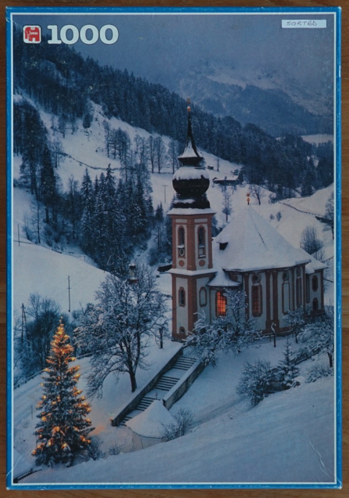 A snow-covered alpine church at twilight with a glowing Christmas tree in the foreground.