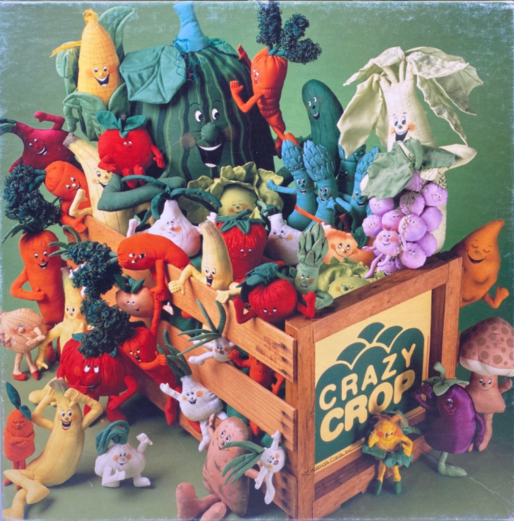 A wooden crate overflowing with anthropomorphic fabric vegetables and fruits with smiling faces against a green background.
