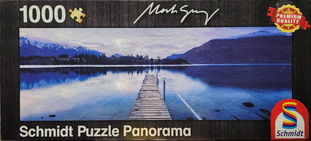 A panoramic landscape photograph of a wooden pier extending into a calm, blue mountain lake at dusk.