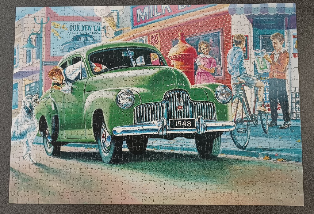 A vintage green Holden car parked on a street in front of a 1940s-style store with children and a dog nearby.
