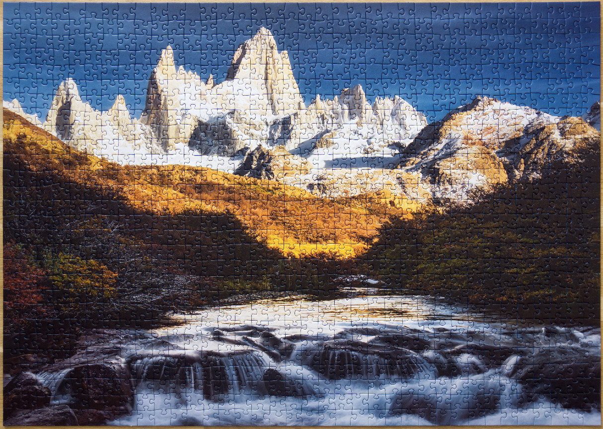 A scenic view of a jagged, snow-capped mountain range above a rushing river and autumn landscape.