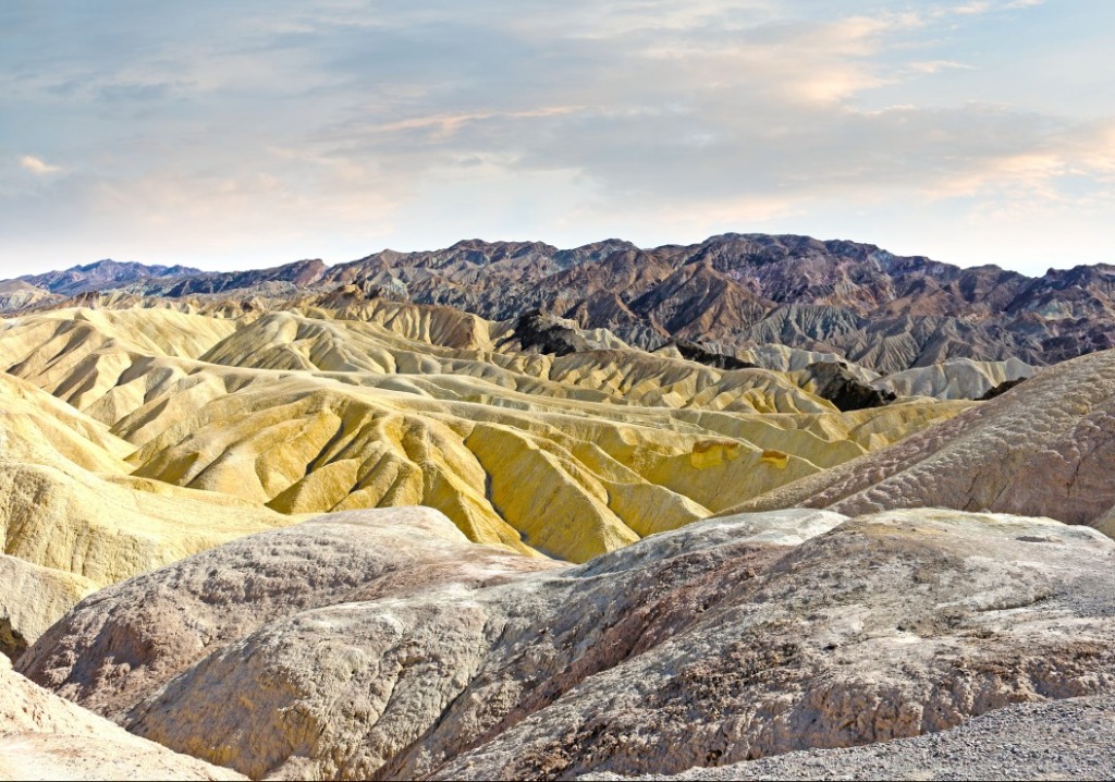 A panoramic view of the golden, eroded badlands and rugged, dark mountain ranges under a soft, cloudy sky.