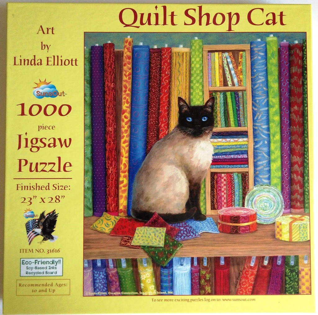 A Siamese cat sitting on a wooden shelf in front of tall, vibrant rolls of colorful patterned quilt fabric.