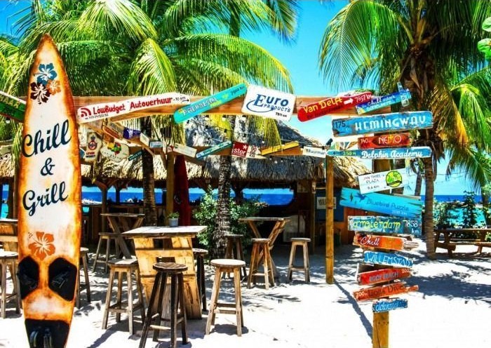 Tropical beach bar with a surfboard sign and numerous wooden directional signposts under palm trees.